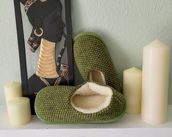 May include: Green knit slippers with a soft, cream-colored lining. The slippers are displayed with a black and gold decorative wall hanging and several ivory candles, creating a cozy, homey scene.