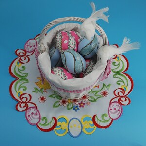 May include: A white wicker basket filled with colourful Easter eggs decorated with floral patterns. The basket is sitting on a white doily with a floral design.