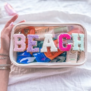May include: A clear, rectangular cosmetic bag with the word "BEACH" spelled out in colorful, textured letters. The bag is filled with various sunscreen bottles and tubes. The bag is held by a person.