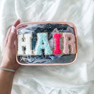 May include: Clear plastic makeup bag with a pink zipper and the word "HAIR" spelled out in colorful fuzzy letters.