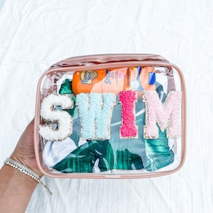 May include: Clear plastic zippered pouch with a pink trim. The pouch contains a white swimsuit with a green tropical leaf print and the word "SWIM" in colorful felt letters. The pouch also contains two bottles of sunscreen.