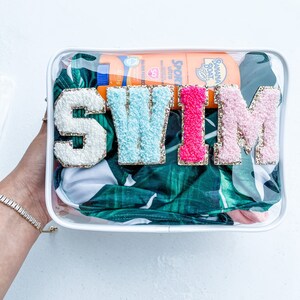 May include: Clear plastic bag with a white zipper closure. The bag contains a swimsuit, sunscreen, and a bottle of sunscreen. The bag also has the word "SWIM" spelled out in large, glittery letters.