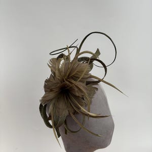 May include: A grey fascinator featuring a decorative arrangement of feathers and fabric. The feathers are in shades of brown and tan, with dark green accents. The base is a rounded shape, mounted on a thin wooden stand.
