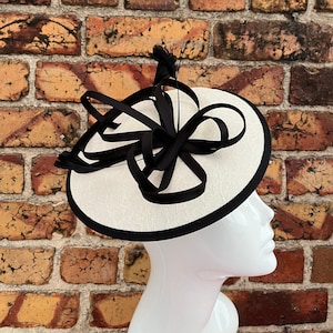 May include: A white hat with a black ribbon and feather accent. The hat has a black trim around the edge.