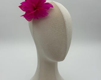 May include: A vibrant fuchsia fascinator headpiece featuring a cluster of soft, layered feathers and a textured, woven base. The accessory is secured to a neutral-toned, smooth, abstract mannequin head display, showcasing the bright pink colour.