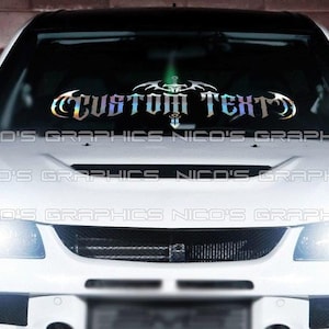 May include: A white car with a black roof and a custom text decal on the windshield. The decal has a decorative design with the words "CUSTOM TEXT" in a holographic, iridescent finish. The car's headlights are on.