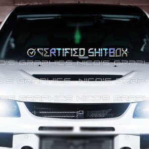 Könnte beinhalten: Ein weißes Auto mit einer schwarzen Windschutzscheibe und einem Aufkleber mit der Aufschrift "CERTIFIED SHITBOX" auf der Windschutzscheibe.