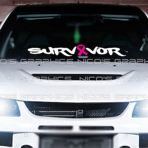 Könnte beinhalten: Weißes Auto mit einem Aufkleber auf der Windschutzscheibe mit der Aufschrift "SURVIVOR" in weißer Schrift, mit einer rosa Schleife, die den Buchstaben "I" ersetzt. Motorhaube und Scheinwerfer des Autos sind sichtbar.