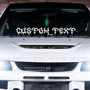 May include: A white car with a black grille and headlights. The windshield has a white vinyl decal that says "CUSTOM TEXT" in a stylized font.