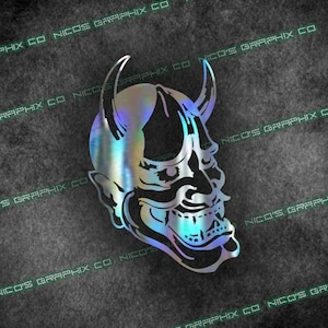 May include: A holographic sticker of a Japanese demon mask with a fierce expression. The mask is black and white with a blue and green iridescent finish.