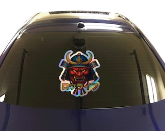 May include: A holographic sticker featuring a red-faced Japanese demon with a blue and gold helmet. The sticker has Japanese text and is displayed on a car window.