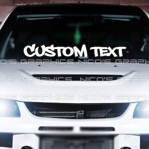 May include: White car with the text "CUSTOM TEXT" in a bold, stylized font across the windshield. The car has bright headlights and a black roof. The text "NICO'S GRAPHICS" is repeated along the bottom.