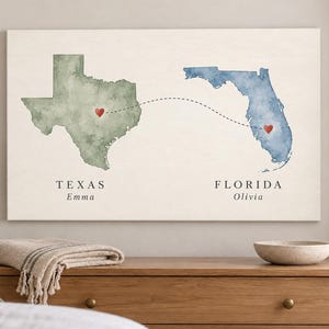 May include: A rectangular wall art print featuring a watercolor map of Texas in green and Florida in blue. A dashed line connects the two states with a red heart on each. The text reads TEXAS Emma and FLORIDA Olivia in a black serif font.