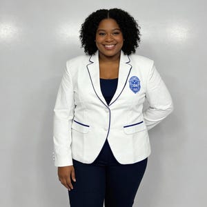 May include: A white blazer with navy blue trim and a crest on the chest. The blazer is paired with navy blue pants. The person is smiling. The blazer has two front pockets and a two-button closure.