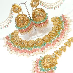 May include: A gold-toned jewelry set featuring earrings, a necklace, and a headpiece. The earrings are large, bell-shaped, and adorned with pink and teal beads. The necklace and headpiece have similar designs, with intricate detailing and pearl accents.
