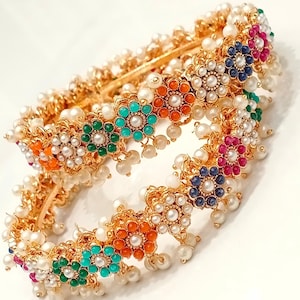 May include: Two gold bangles with white pearls and colorful flower-shaped accents. The flowers have a gold center and are surrounded by small white pearls.