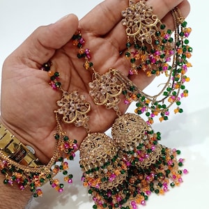 May include: A pair of gold-toned jhumka earrings featuring intricate floral-shaped studs adorned with clear crystals. The earrings are embellished with dangling chains and clusters of small green, pink, and orange beads that create a layered effect.