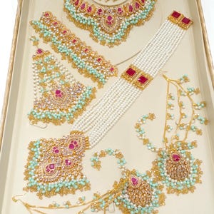 May include: An assortment of gold-toned jewelry sets, including necklaces, earrings, and headpieces, displayed in a beige tray. The jewelry features red gemstones, white pearls, and turquoise-colored accents. The pieces are intricately designed with detailed patterns.