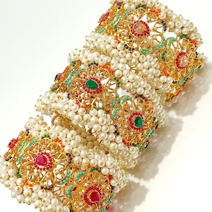 May include: Ornate gold bangles with intricate floral designs, adorned with white pearls and colorful gemstones. The bangles feature red, green, orange, and turquoise accents, creating a vibrant and luxurious look. The jewelry is displayed on a white surface.