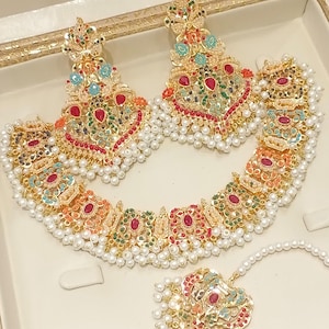 Può includere: Un set di gioielli color oro composto da collana, orecchini e copricapo. I pezzi sono adornati con pietre preziose colorate, tra cui rosso, verde e blu, e rifiniti con piccole perle bianche. Il set è esposto in una scatola color crema.