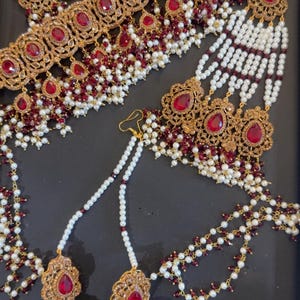 May include: A gold-toned jewelry set featuring a wide choker necklace, a pair of jhumka earrings, and a maang tikka. The pieces are adorned with teardrop-shaped red stones, small white pearls, and tiny dark red beads in an intricate metal design.