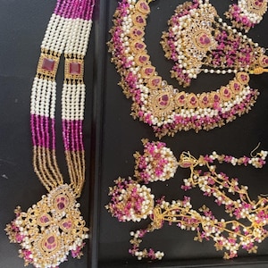 May include: An ornate jewelry set featuring a necklace, earrings, and a headpiece. The pieces are gold-toned with pink and white accents, including beads and gemstones. The set is displayed on a black surface.