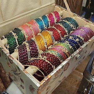 May include: A rectangular jewelry box with a floral pattern containing rows of multi-colored glittery bangles. The bangles are arranged in shades of green, pink, blue, yellow, red, purple, and dark red, each featuring a small gold-toned charm.