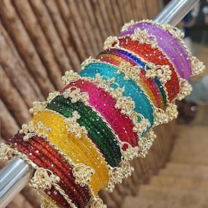May include: A selection of vibrant bangles in red, yellow, green, blue, pink, and purple. Each bangle is decorated with gold-coloured floral details. The bangles are displayed on a silver rod, highlighting their bright colours.