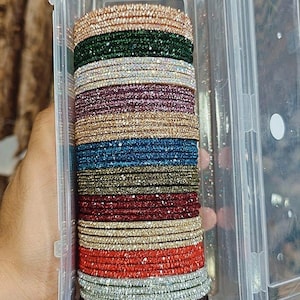 Può includere: Un contenitore di plastica trasparente pieno di pile di braccialetti colorati e glitterati. I braccialetti sono in vari colori, tra cui oro, verde, rosso, blu e argento. Il contenitore è tenuto in una mano.