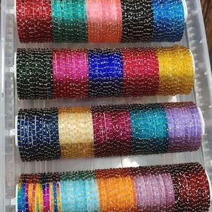 May include: A collection of thin, circular bangles arranged in four horizontal rows within a clear plastic container. Each bangle features a textured, glittery surface in various vibrant colours including red, green, orange, black, blue, yellow, pink, and purple.