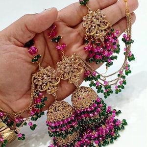 May include: A pair of gold-tone jhumka earrings featuring intricate floral metalwork, clear crystals, and cascading strands of small pink and green beads. The design includes a chain attachment with additional bead accents and a bell-shaped base.
