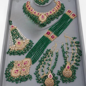 May include: A detailed jewellery set comprising a necklace, bracelet, earrings, and a headpiece. The set features gold-toned metalwork with ruby-coloured accents and green beaded strands. The pieces are displayed on a grey surface.