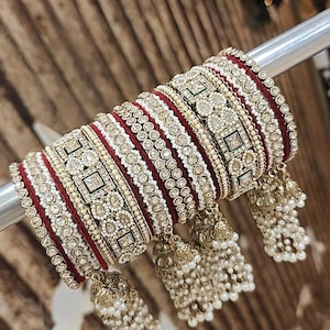 Op de afbeelding: Een verzameling sierlijke armbanden met een rood en goud kleurenschema. De armbanden hebben ingewikkelde details met witte en gouden versieringen, waaronder kleine, bungelende parelaccenten. De sieraden worden op een zilveren staaf getoond.