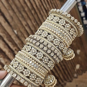 May include: A stack of gold bangles with pearl and rhinestone embellishments. The bangles are decorated with small, mirrored accents and have dangling charms.