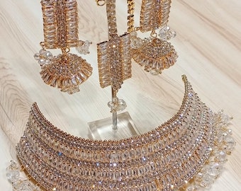 May include: A gold necklace with a multi-layered design featuring clear rhinestones. The necklace has a matching set of earrings and a small pendant.