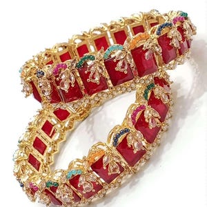 May include: A pair of gold bangles featuring red, square-shaped stones and colourful accents. The bangles are adorned with white pearl-like beads and intricate floral designs.