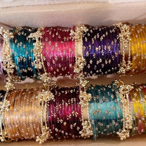 May include: A collection of colorful bangles in a box. The bangles are in various colors, including purple, teal, pink, and gold. Each bangle is adorned with small, decorative beads and gold-colored accents. The bangles are arranged in rows.