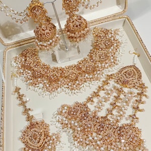 May include: A gold and pearl bridal jewelry set with a necklace, earrings, a maang tikka, and a nose ring. The jewelry is decorated with intricate designs and features a delicate pink hue.