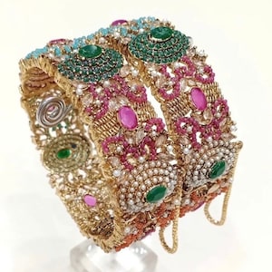 May include: Two ornate gold bangles, intricately designed with a variety of gemstones. The bangles feature emerald, ruby, and pearl accents, with a filigree pattern. The overall design is detailed and luxurious, suitable for special occasions.
