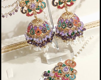 May include: Ornate gold-toned earrings and a matching headpiece. The earrings feature a floral design with colorful gemstones and dangling pearl strands. The headpiece mirrors the earring design, with a similar arrangement of gems and pearls. The jewelry is displayed on a clear stand.