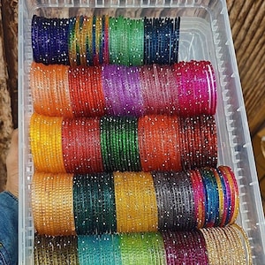 May include: A clear plastic container filled with stacks of colorful bangles. The bangles are in various colors, including blue, green, orange, red, purple, yellow, and gold. Each bangle is adorned with small, sparkling embellishments.