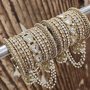 May include: Gold-toned bangles with intricate detailing and embedded clear, triangular-shaped stones. Some bangles have dangling pearl accents. The jewelry is displayed on a metal rod against a blurred wooden background.