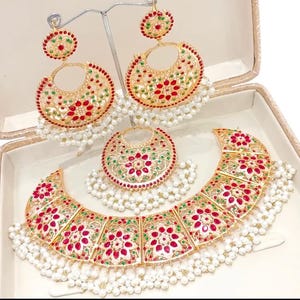 May include: A gold-tone necklace and earrings set with red, green, and white stones. The necklace has a crescent moon shape with a floral design and is adorned with pearl drops. The earrings are also crescent moon shaped with floral designs and pearl drops.