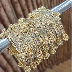 Könnte beinhalten: Eine Sammlung goldfarbener Armreifen, jeder mit kleinen, funkelnden Steinen besetzt. Die Armreifen sind mit dekorativen floralen und perlenbesetzten Akzenten verziert. Der Schmuck wird auf einer silbernen Stange vor einem verschwommenen braunen Hintergrund präsentiert.