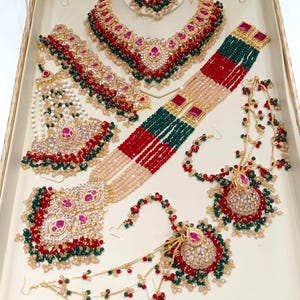 May include: An assortment of ornate jewelry pieces, including necklaces, earrings, and bracelets, displayed in a white box. The jewelry features red, green, and gold accents, with a variety of bead and stone embellishments. The pieces are arranged to showcase their intricate designs.