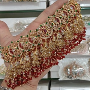 May include: An elaborate gold necklace with red, green, and clear gemstones. The necklace has a detailed design with dangling elements and a beaded fringe. The piece is ornate and suitable for formal events or celebrations.