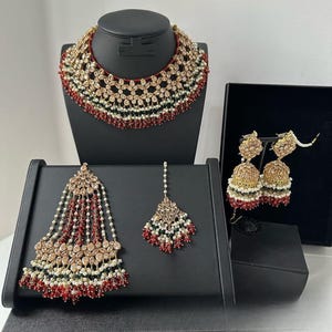 May include: An ornate jewelry set featuring a necklace, earrings, and headpieces. The necklace is displayed on a black mannequin head. The jewelry features gold-toned metal, red, white, and green beads, and clear gemstones. The set is displayed on black stands.