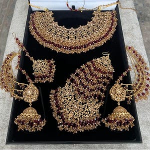 Pode incluir: Um conjunto completo de joias douradas com intrincados desenhos florais e detalhes em bordô. O conjunto inclui um colar, brincos, uma peça de cabeça e outras peças combinando, tudo exibido em uma caixa preta. As joias são ornamentadas.