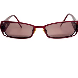 May include: Red rectangular sunglasses with a thin metal frame and tinted lenses. The temples have a patterned design. The sunglasses have a modern, geometric design.