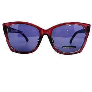 May include: Red cat-eye sunglasses with a dark purple lens and the brand name "BCBGMAXAZRIA" printed on the right lens.